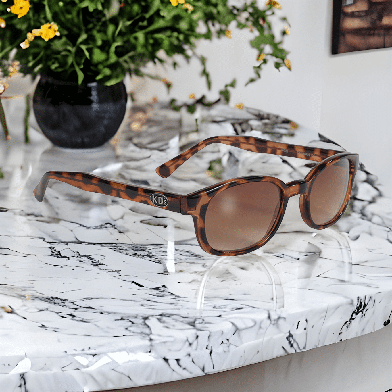 KD's 200 Sonnenbrille mit bernsteinfarbenen Gläsern und einem verzierten Rahmen in Schildpattoptik, passend für Motorradfahrer und Sportler auf einer weißen Marmortheke vor einer Pflanze.